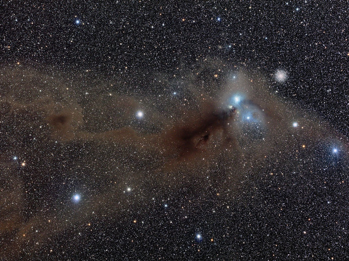This spectacular wide field image shows the area around the star R Coronae Australis. A huge dust cloud, about eight light-years across, dominates the centre of the image. The bluish reflection nebula close to R Coronae Australis is right of centre and the globular cluster NGC 6723 lies to the upper-right of the nebula. Corona Australis is a tiny tiara-shaped constellation, located next to the larger constellation of Sagittarius, in the direction of the centre of the Milky Way. In spite of its faintness, this southern winter constellation can be easily spotted from dark sites because of its characteristic shape and position in the sky.